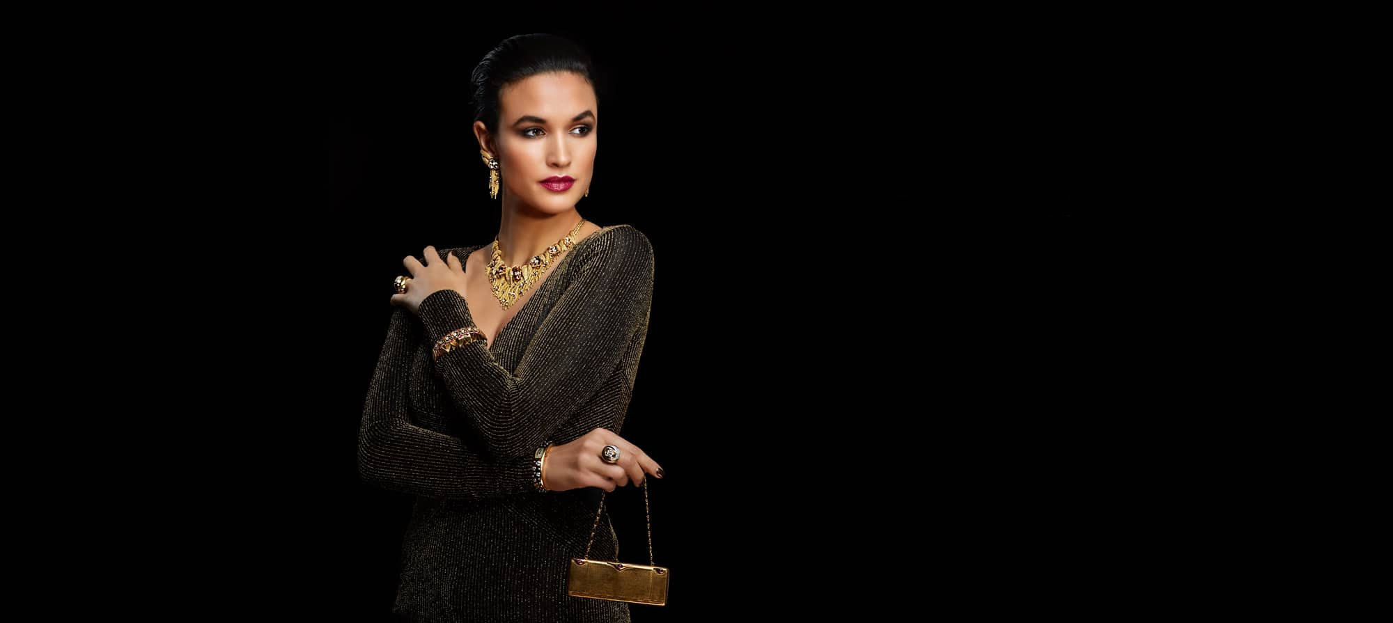 Woman facing forward staring left wearing a gray dress holding a small gold clutch purse with matching gold necklace and earrings, two bracelets and two rings with a black background