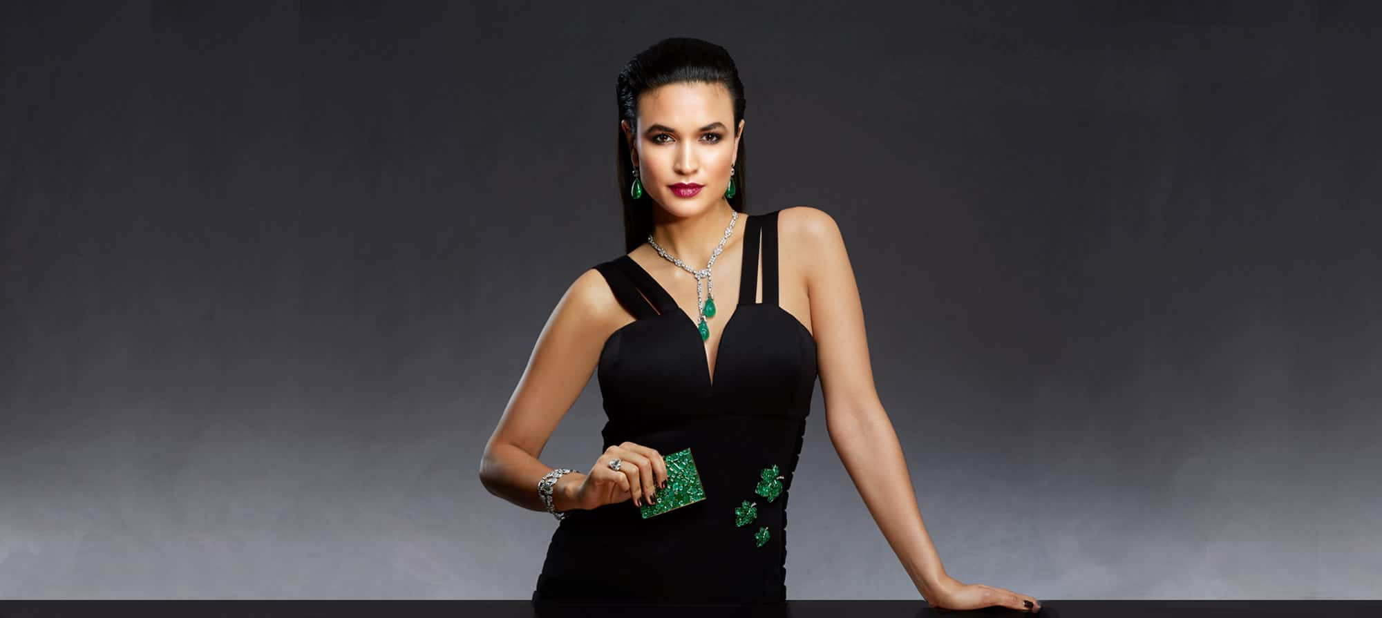 Women wearing black dress with matching teardrop emerald earrings and necklace she is holding a green piece of material, gray dark background