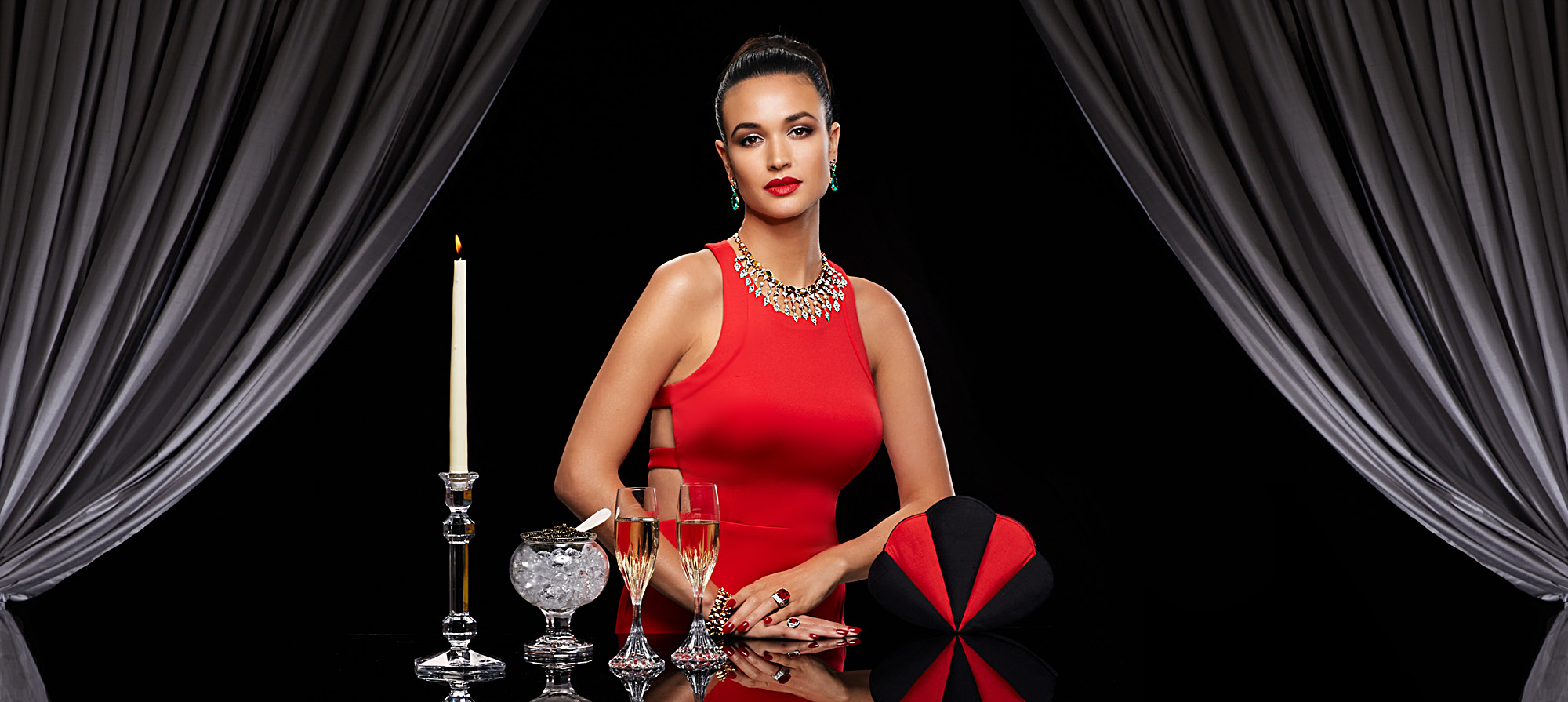 Woman looking forward sitting at a table with two glasses of champagne, ice and a single candle wearing red dress with emerald earrings, diamond necklace and ruby rings dark black background