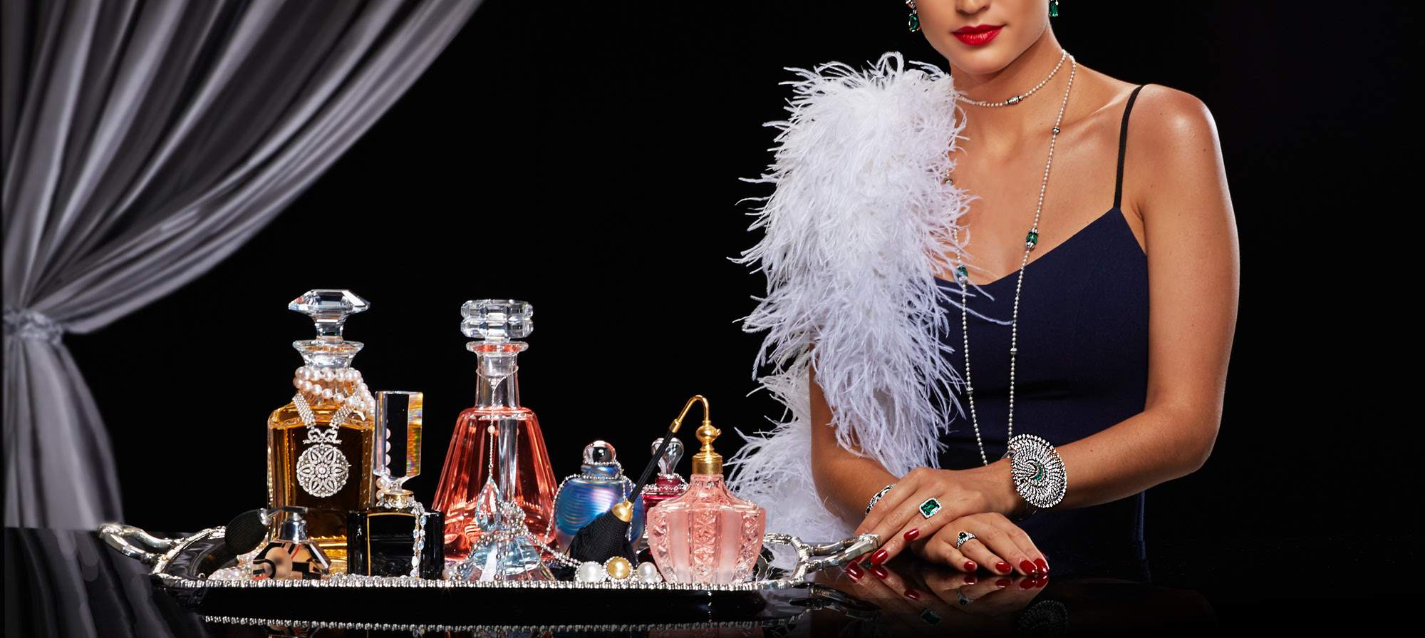 Woman wearing a black dress with a tray of perfumes sitting next to her, wearing a white feather boa and vintage antique emerald earrings, necklace, bracelet and rings black background