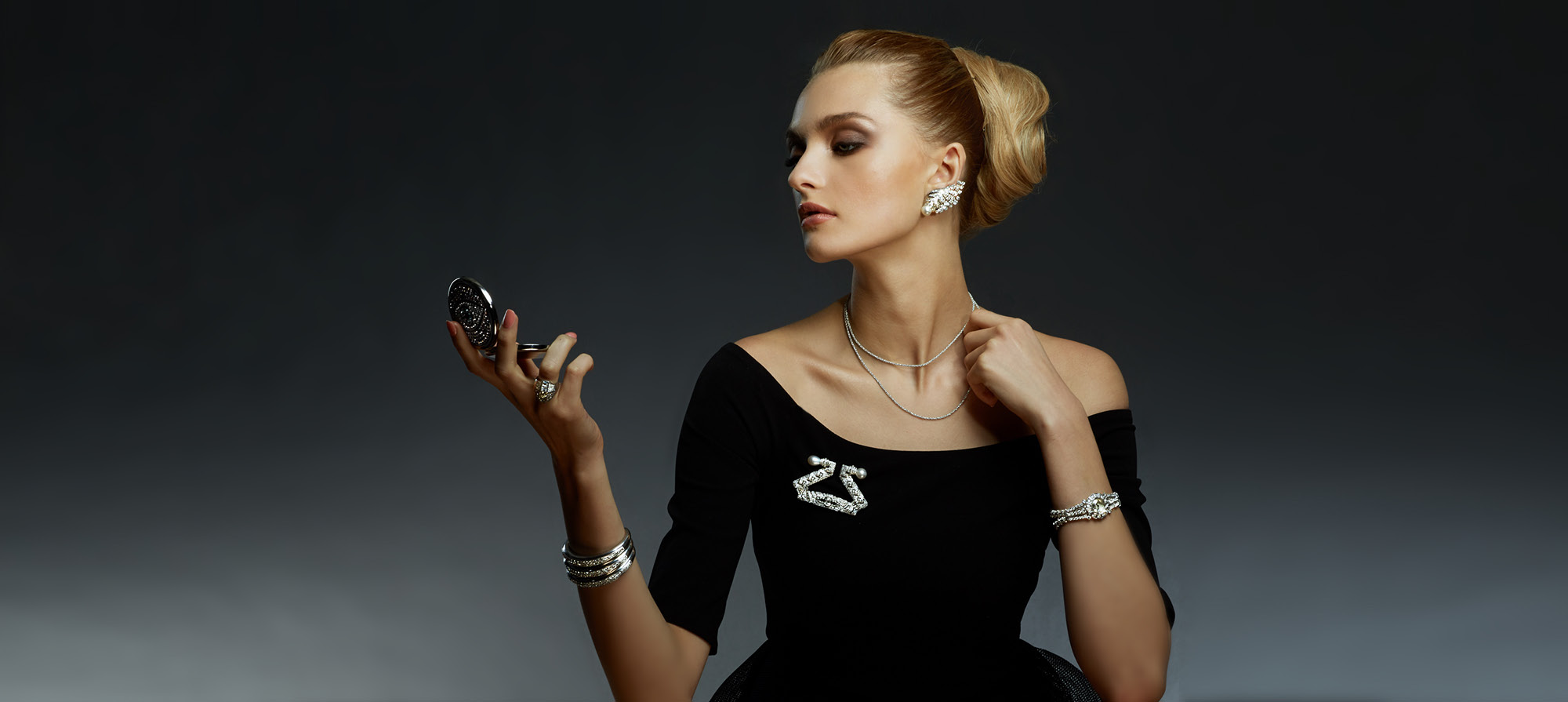 Woman wearing black dress looking at a makeup compact mirror wearing vintage antique earrings, necklace, broach, bracelets and rings dark gray black background